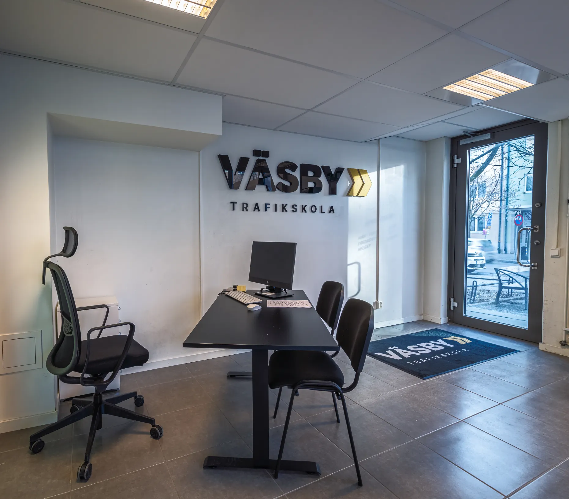 En modern kontorsinställning med en dator på ett svart bord; tre stolar omger den. På väggen står det "VÄSBY TRAFIKSKOLA" och ett fönster visar en urban gatuvy.